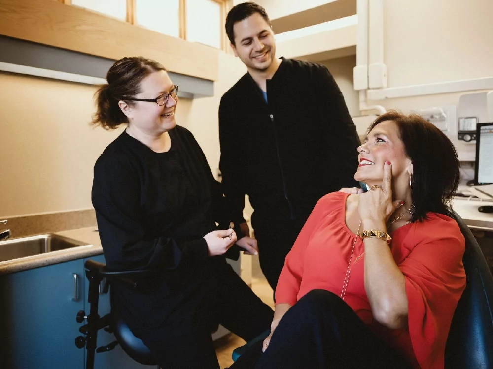 The image shows three individuals in a room, which appears to be an office or medical setting. One person is seated by a sink, another is standing wearing a black jacket, and the third is seated near a computer in a red blouse. The room features beige walls and wooden cabinets.