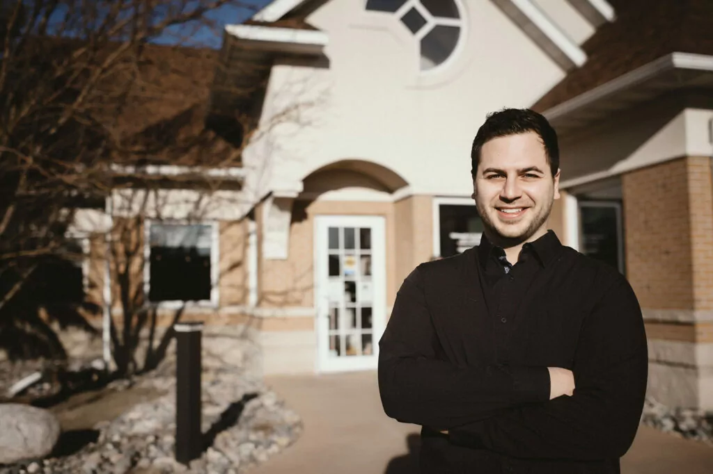 Dr. Majidian in front of his dental office in Fargo, nd, with brick accents and a white glass-paneled door. The setting includes a circular window above the door, manicured landscaping, and a bright blue sky, conveying a professional, welcoming environment.