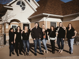 Dental team of Majidian Dental black tops and jeans stand arm-in-arm in front of a brick-and-stucco building with a steep roof. The setting includes a paved area with landscaping and architectural features like a circular window and arched entryway.